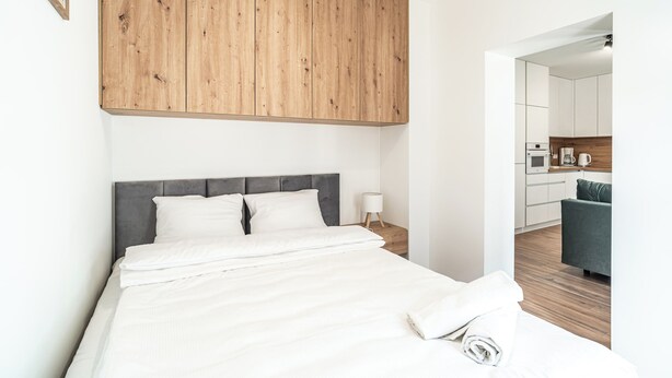 Una habitación de hotel moderna con una cama grande, un panel de pared de madera, un cabecero gris, una mesita de noche con lámpara y vistas al área de la cocina.