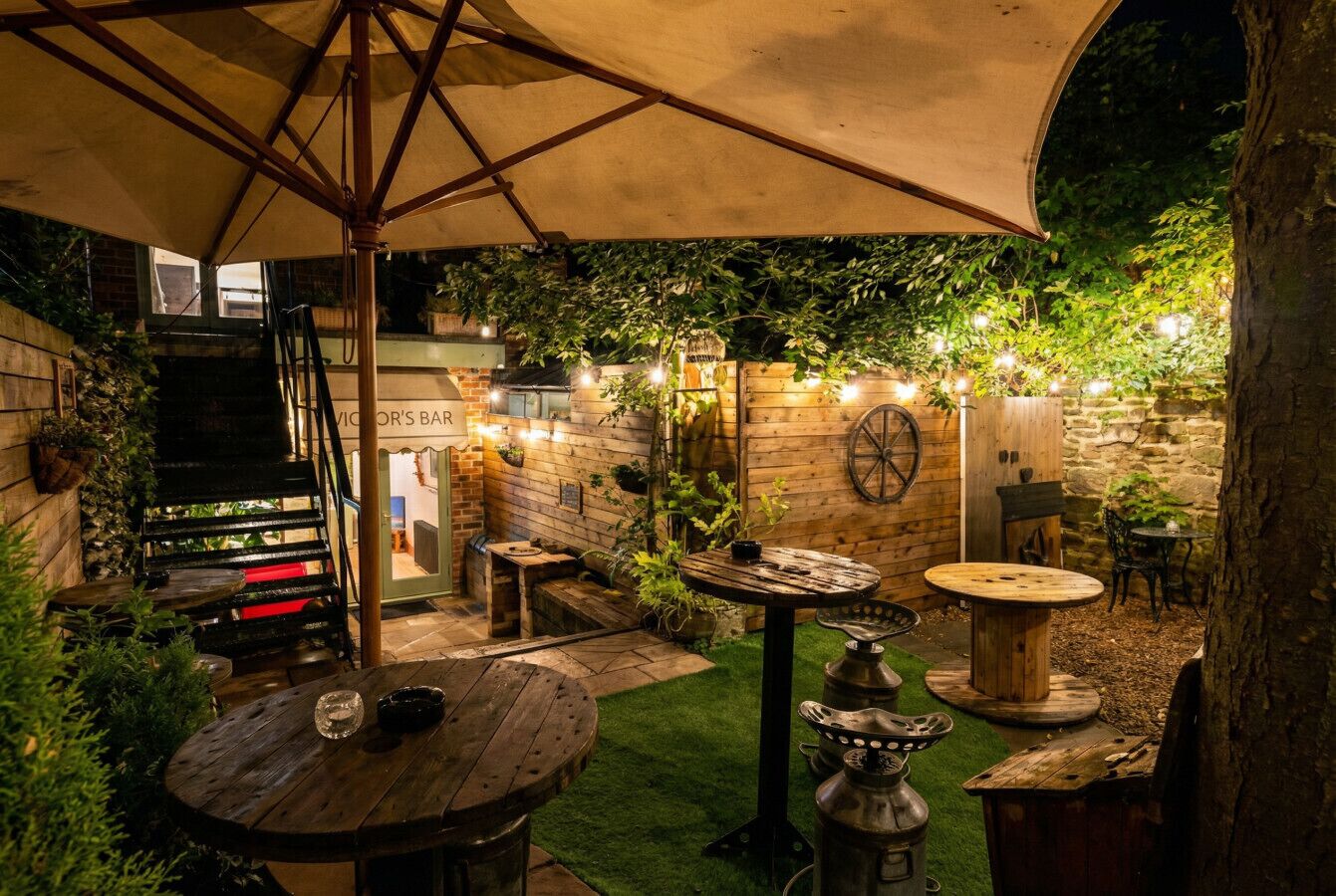 An outdoor patio area with wooden tables and chairs, a large umbrella, and string lights.