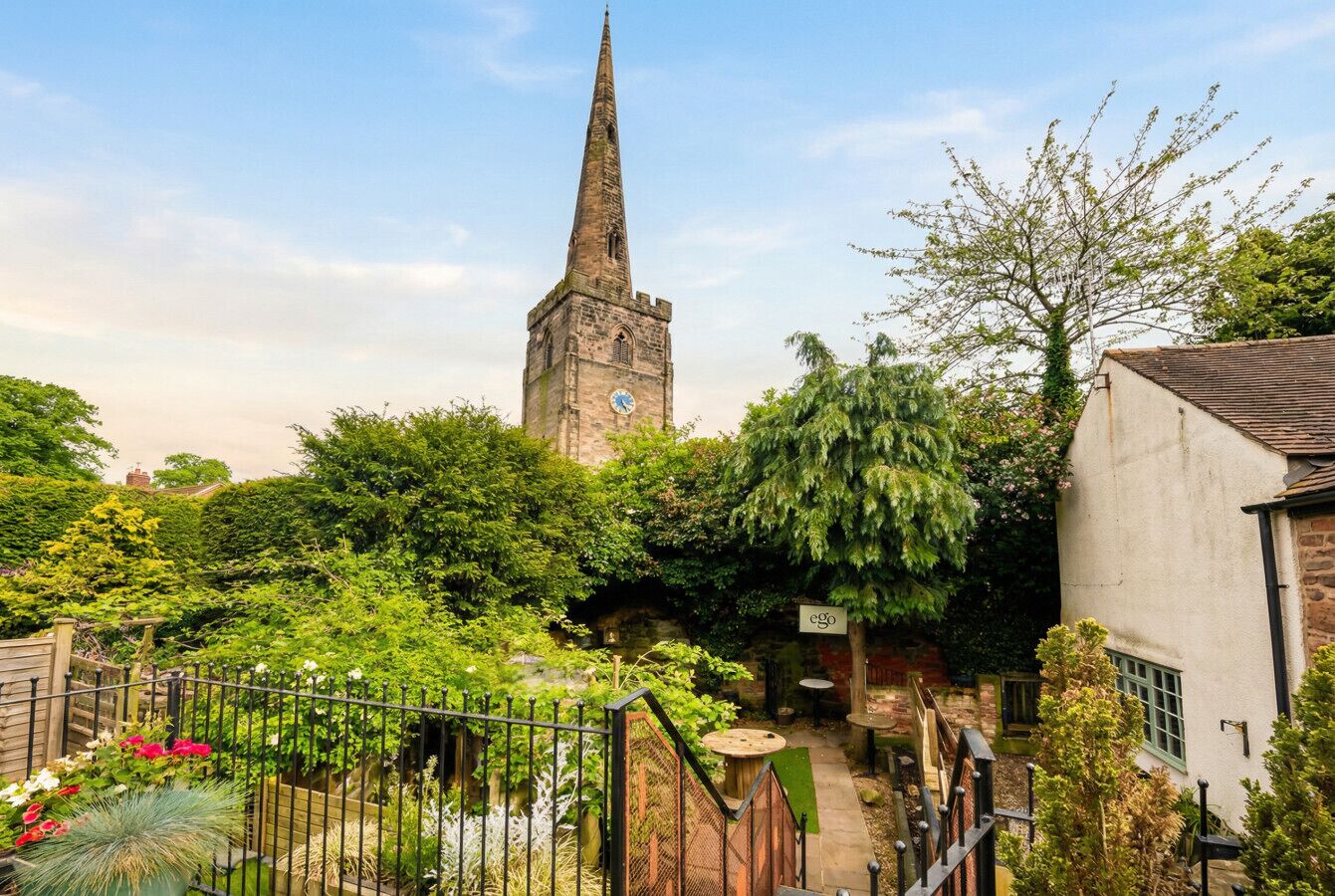 A church with a tall spire, surrounded by lush greenery and a small garden area.
