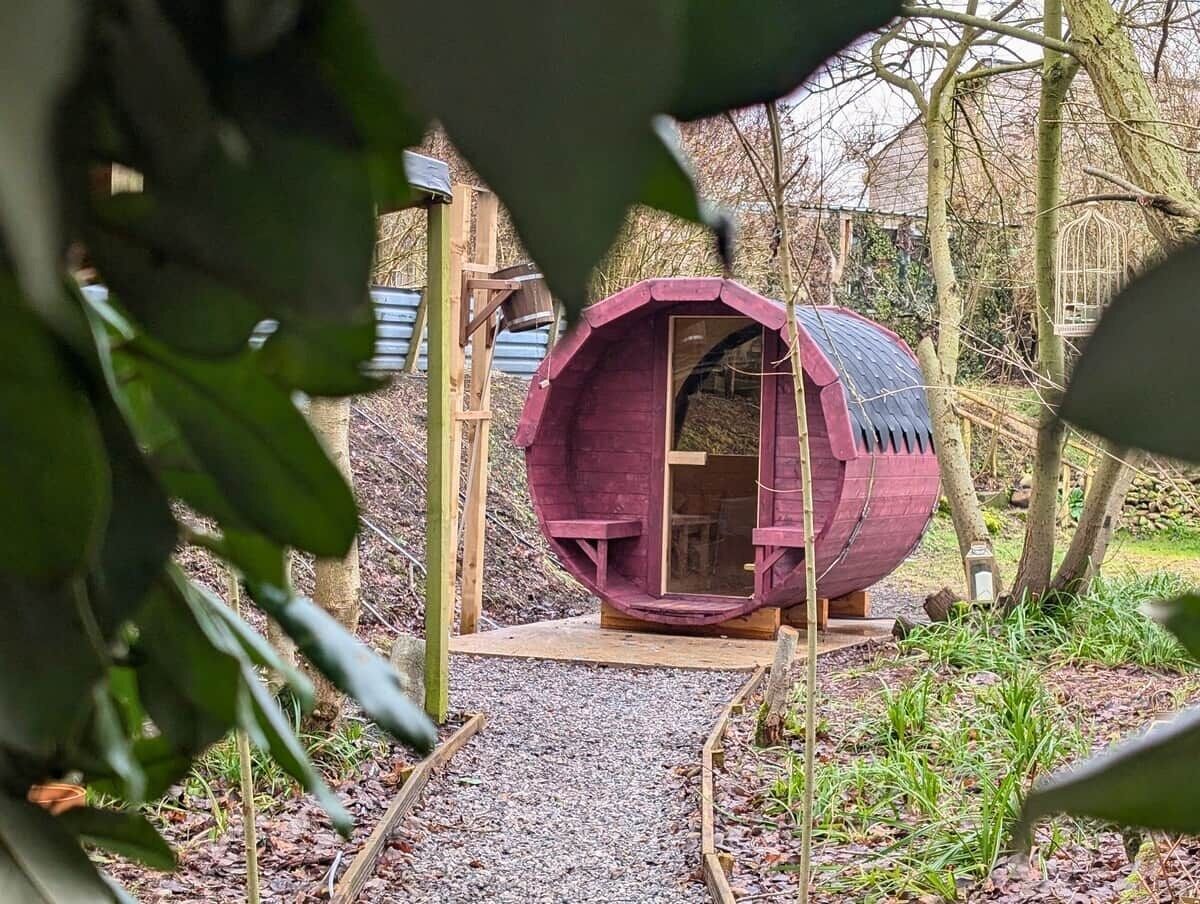 A cylindrical wooden structure with a door, surrounded by trees and gravel.