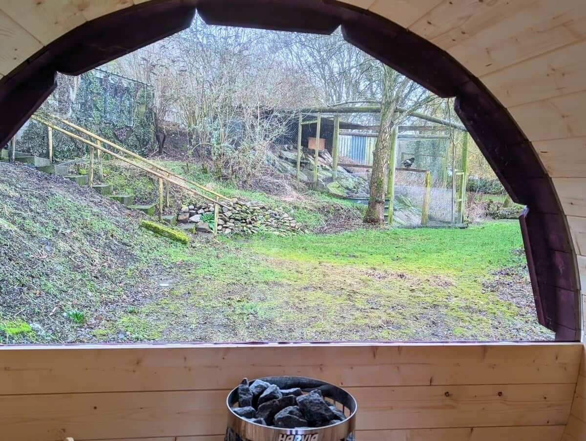 A view from a wooden room with a window, a metal container on the floor, and a grassy area with a wooden structure in the background.
