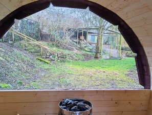 Blick von einem Holzraum mit Fenster, einem Metallbehälter auf dem Boden und einer grünen Fläche mit einer Holzhütte im Hintergrund.