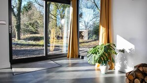 Une pièce dotée de grandes portes-fenêtres coulissantes, d’une plante en pot, d’un vase blanc et d’une pile de bois de chauffage.