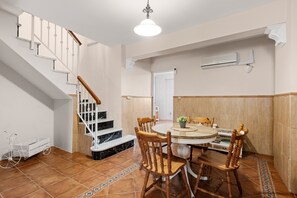 A dining area with a round table, wooden chairs, a staircase, and a white wall with an air conditioning unit.