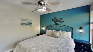 A bedroom with a bed, a ceiling fan, a wall-mounted light fixture, and a painting. - Spring Tide | Pet-Friendly, Large Deck, Great Location (Tybee Island)