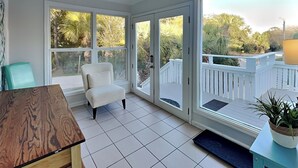 A sunlit room with a white chair, a wooden table, and large windows offering a view of a white deck and greenery. - Spring Tide | Pet-Friendly, Large Deck, Great Location (Tybee Island)