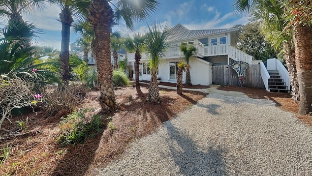 A two-story house with a gravel driveway, palm trees, and a wooden fence. - Spring Tide | Pet-Friendly, Large Deck, Great Location (Tybee Island)