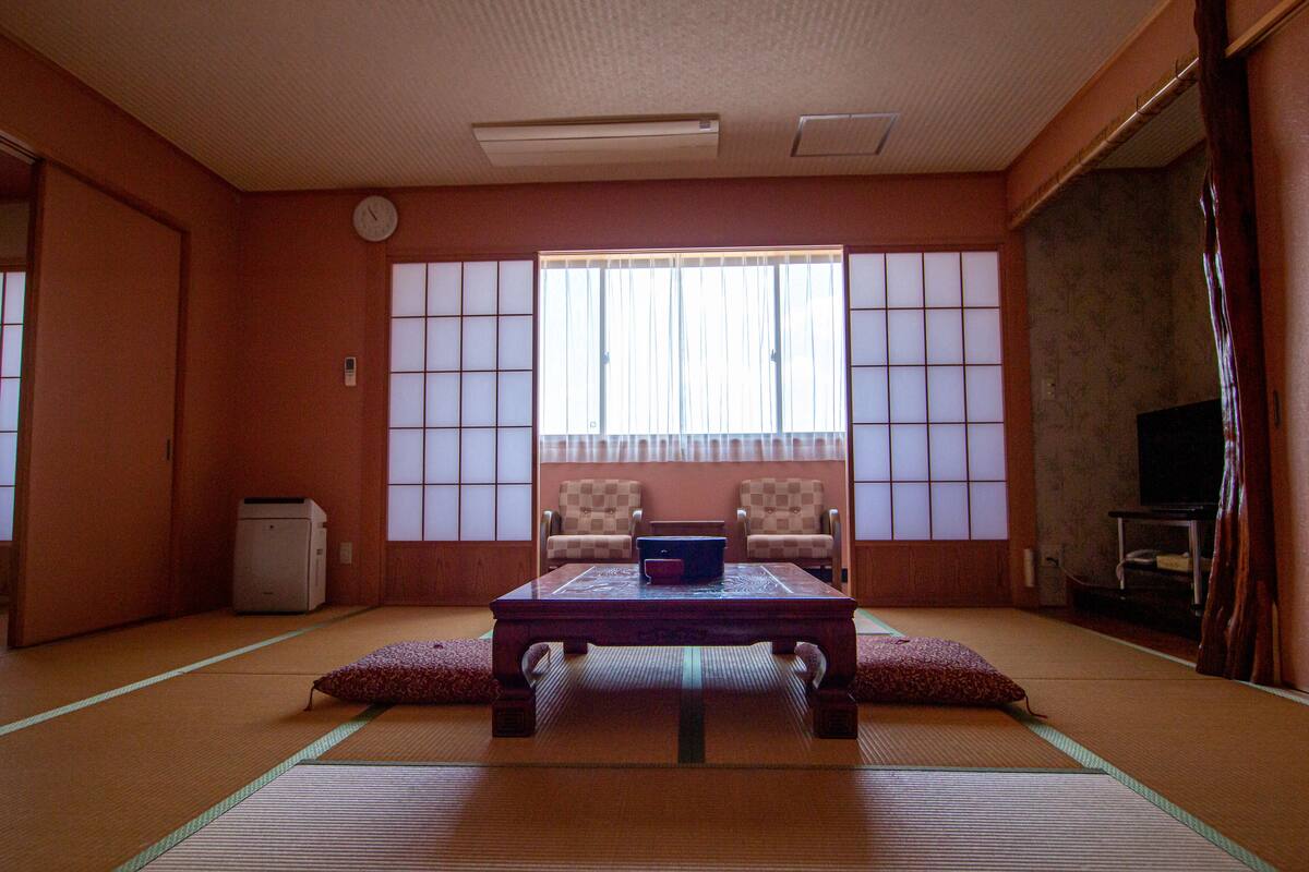 Ett traditionellt japanskt rum med tatamimattor på golvet, ett lågt bord med kuddar, två stolar, en tv och en skjutdörr.
