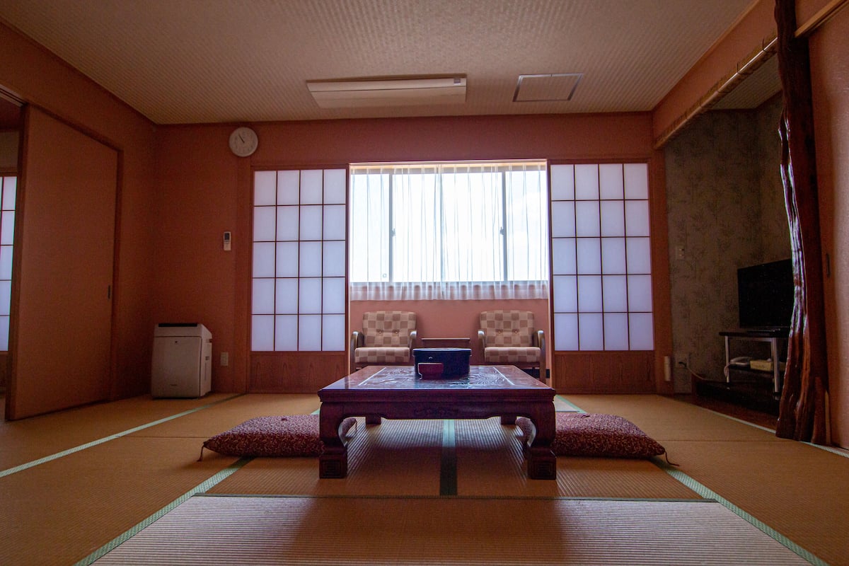 A traditional Japanese room with tatami flooring, a low table with cushions, two chairs, a television, and a sliding door.