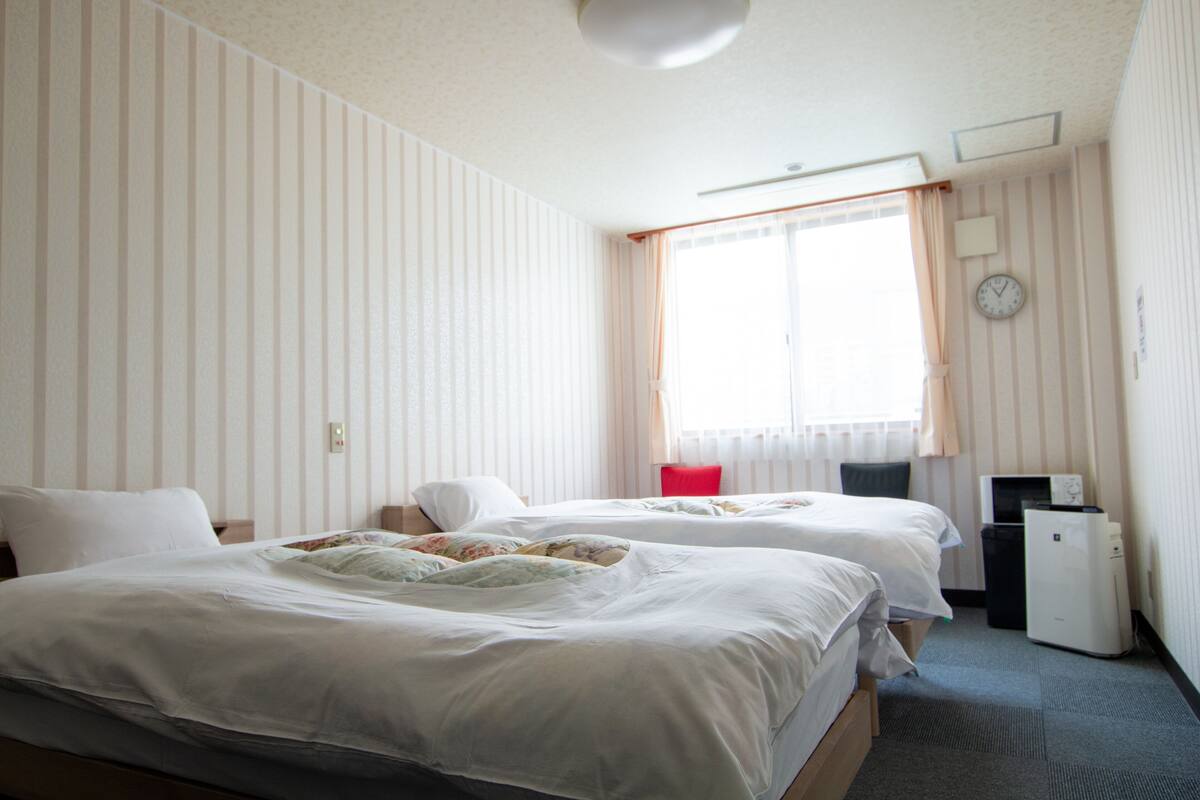 Ett hotellrum med två sängar, ett skrivbord, en stol, en väckarklocka och ett fönster med gardiner.