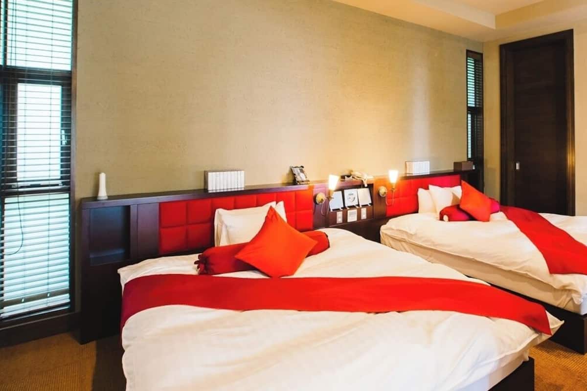 Ein Hotelzimmer mit zwei Betten, einem roten Kopfteil, einem Nachttisch mit Lampe und einem Fenster mit Jalousien.