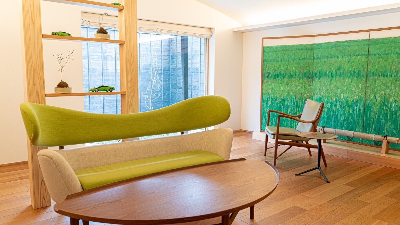 Ein Zimmer mit Holzboden, einem grünen Sofa, einem Holztisch, einem Stuhl und einem großen Fenster mit Blick auf eine grüne Landschaft.