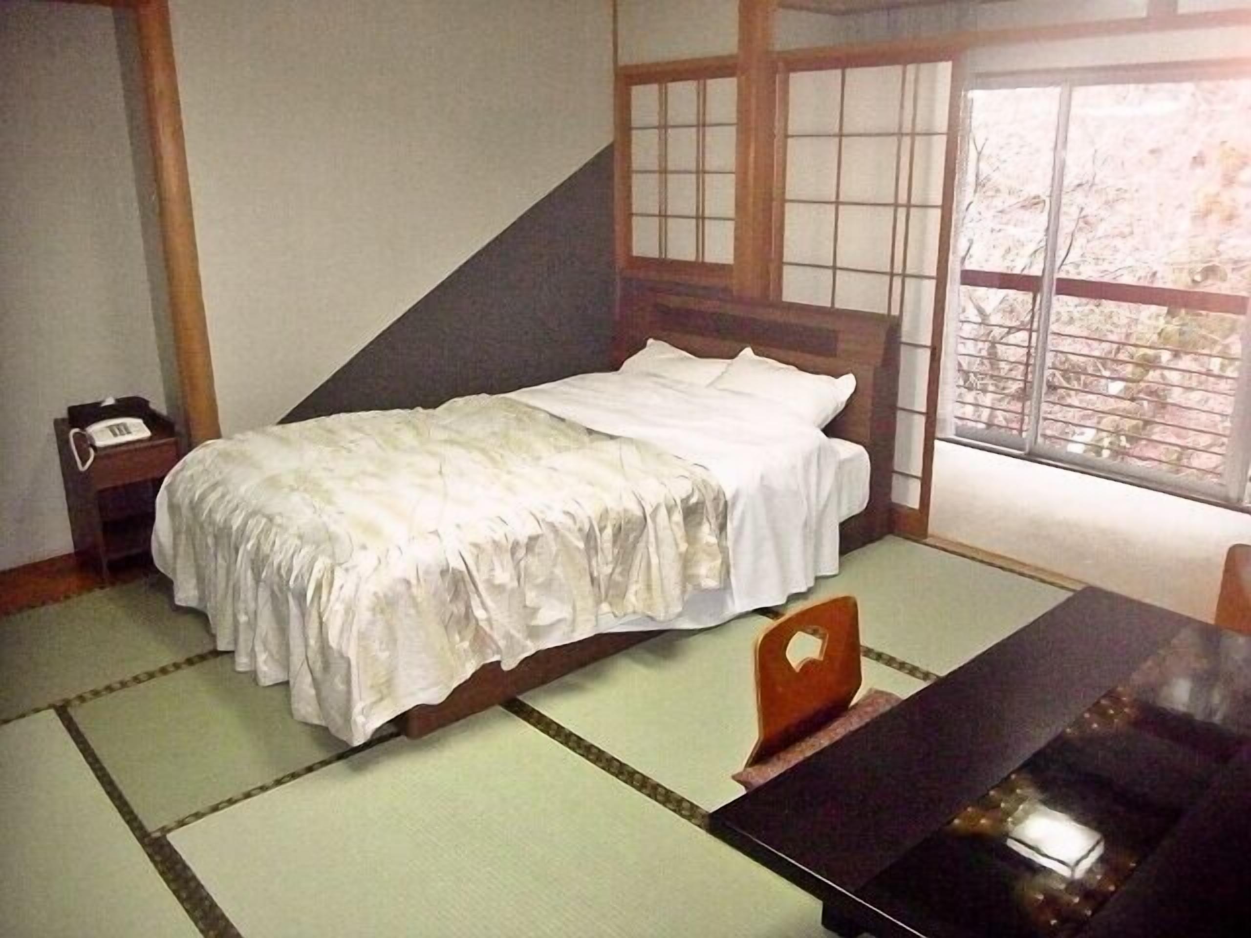 Ein traditioneller japanischer Raum mit Tatami-Böden, einem niedrigen hölzernen Bett, einem kleinen Nachttisch mit Telefon und einem Fenster mit Blick auf Bäume.