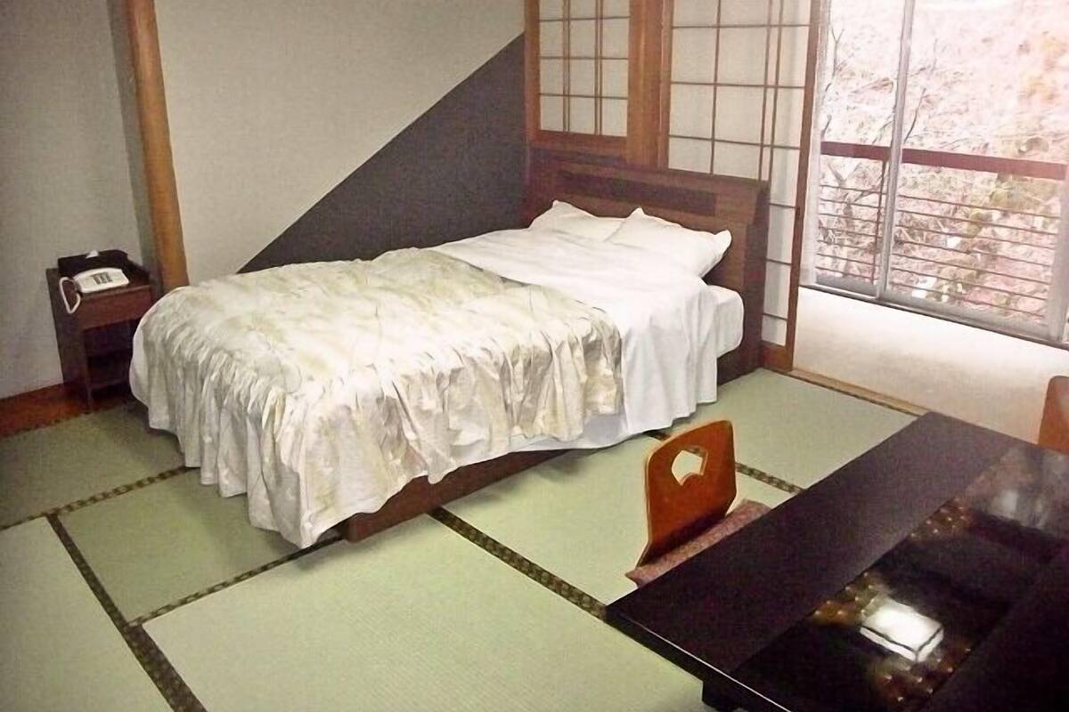 A traditional Japanese room with tatami flooring, a low wooden bed, a small bedside table with a telephone, and a window with a view of trees.