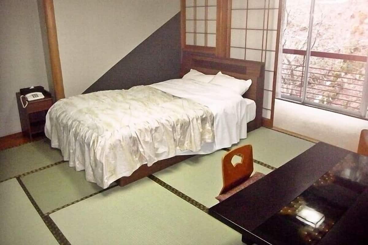 Ein traditioneller japanischer Raum mit Tatami-Böden, einem niedrigen hölzernen Bett, einem kleinen Nachttisch mit Telefon und einem Fenster mit Blick auf Bäume.