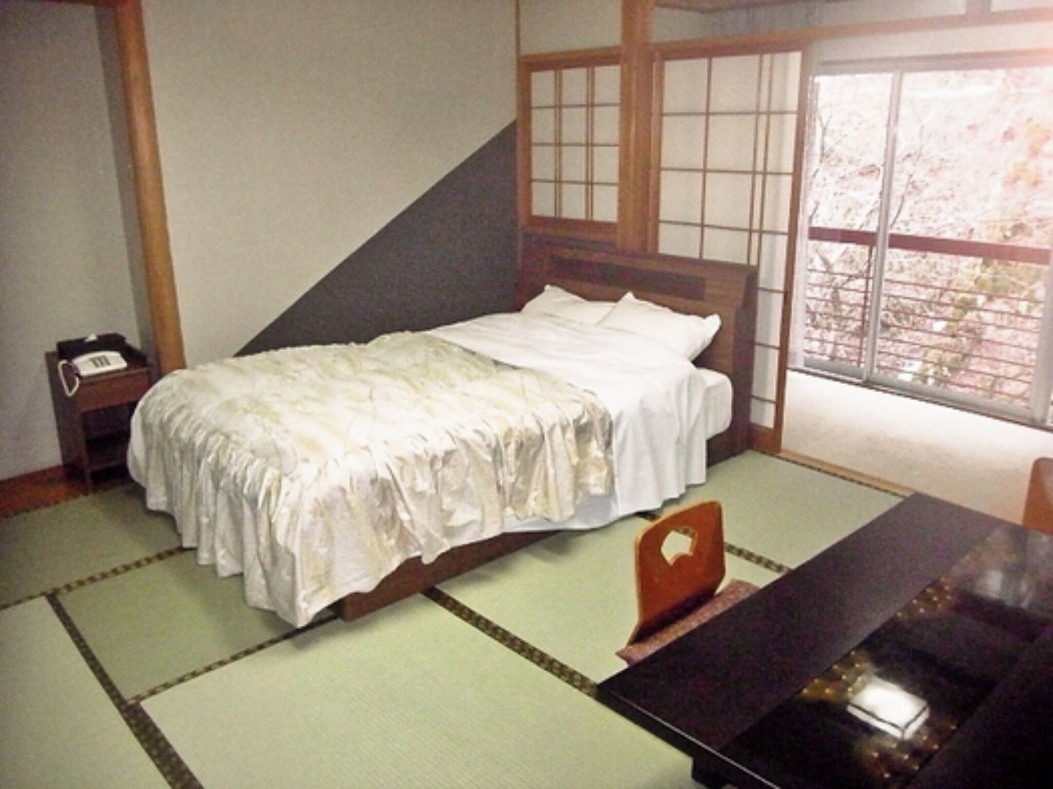 Ein traditionelles japanisches Zimmer mit Tatami-Bodenbelag, einem niedrigen hölzernen Bett mit weißer Bettwäsche, einem kleinen Nachttisch mit Telefon und einer Schiebetür mit Blick ins Freie.
