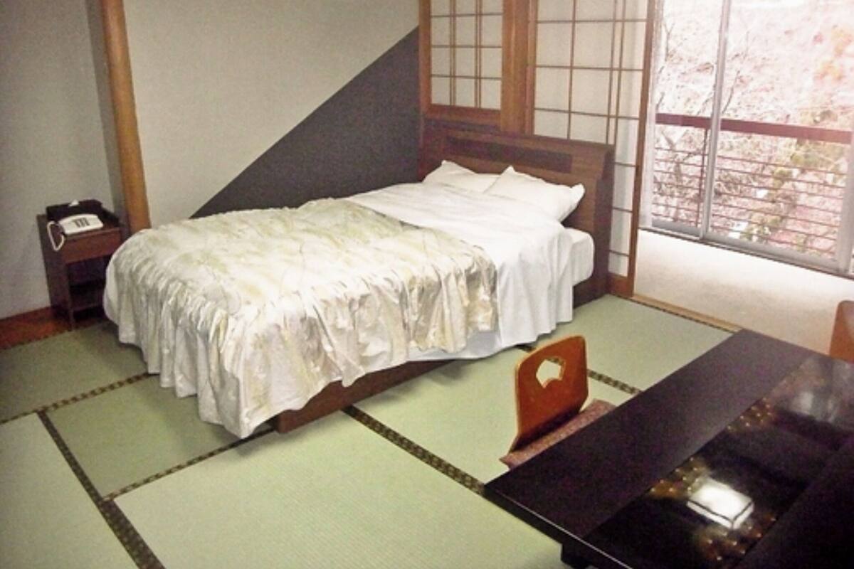 A traditional Japanese-style room with tatami flooring, a low wooden bed with white bedding, a small bedside table with a telephone, and a sliding door with a view of the outside.