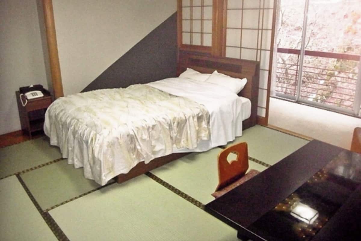 Ein traditionelles japanisches Zimmer mit Tatami-Bodenbelag, einem niedrigen hölzernen Bett mit weißer Bettwäsche, einem kleinen Nachttisch mit Telefon und einer Schiebetür mit Blick ins Freie.