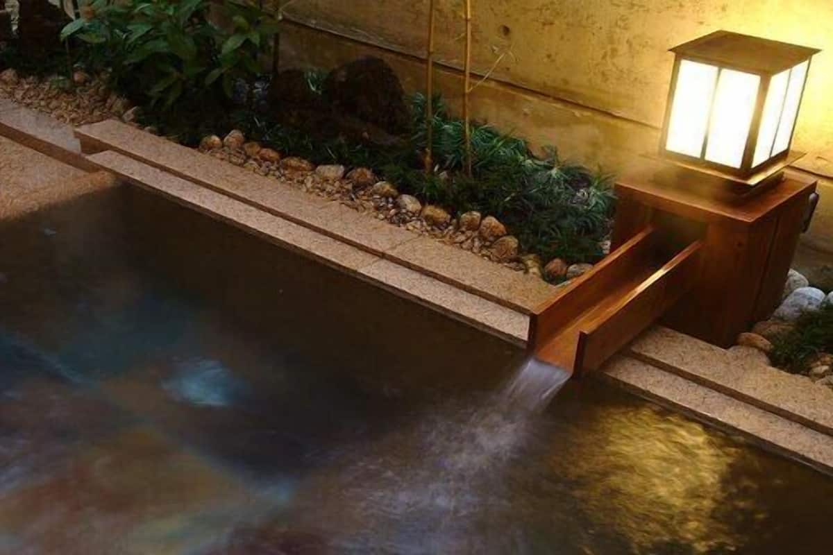 A small indoor pool with a wooden edge and a stone border, a wooden bench, and a wall-mounted lamp.