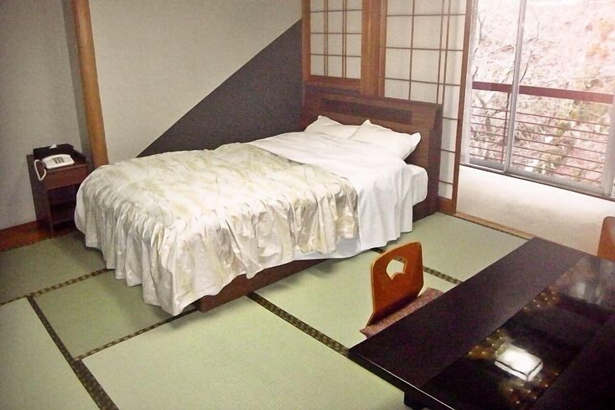 A traditional Japanese room with tatami flooring, a low wooden bed, a small bedside table with a telephone, and a window with a view of trees.