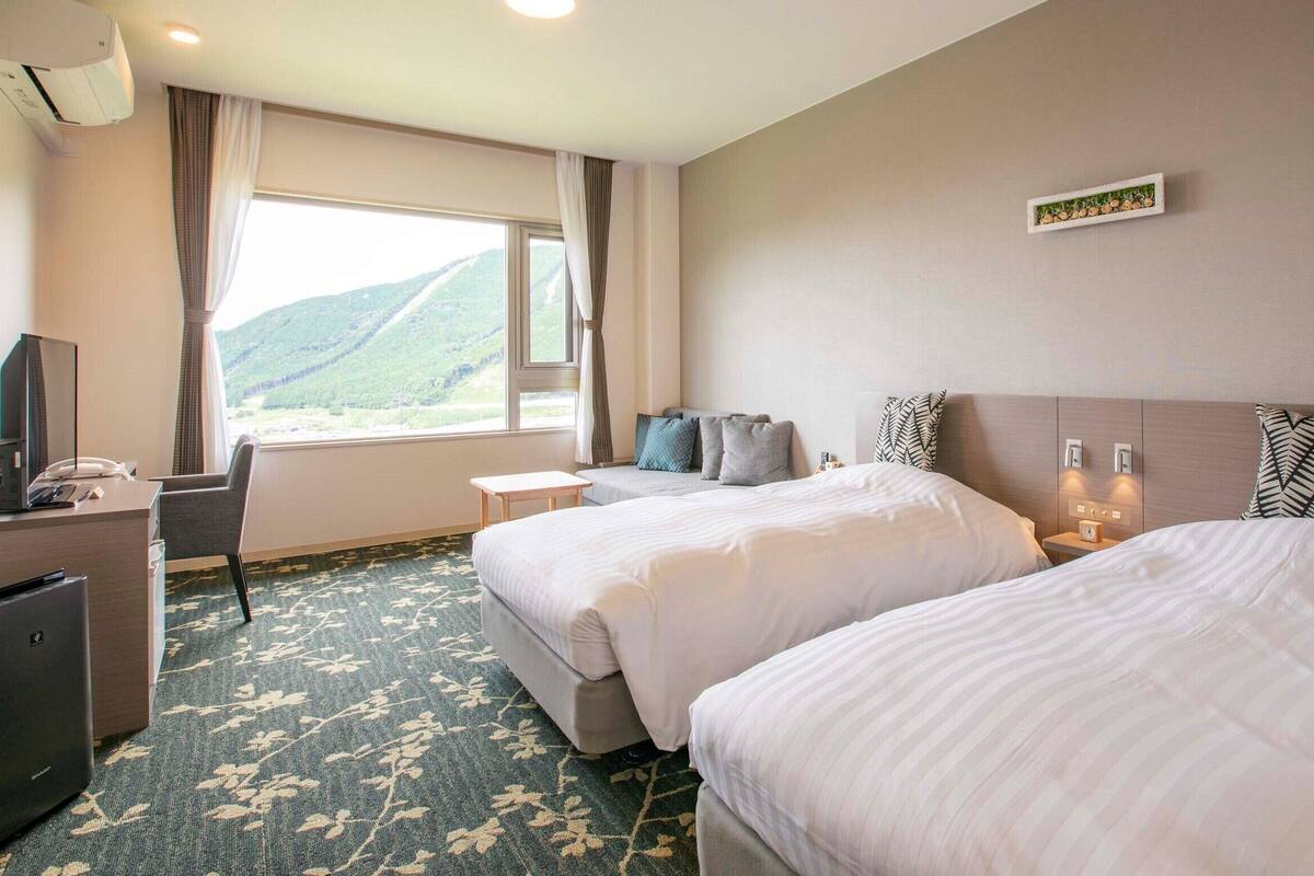 Une chambre d’hôtel avec deux lits, un bureau, une chaise, une télévision et une fenêtre donnant sur une vue sur la montagne.