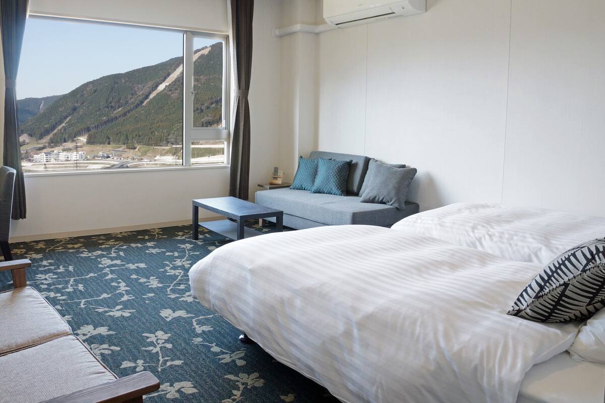 Une chambre d’hôtel comprenant un lit, un bureau, une chaise, une petite table et un canapé. Il y a une fenêtre donnant sur une montagne et un immeuble.