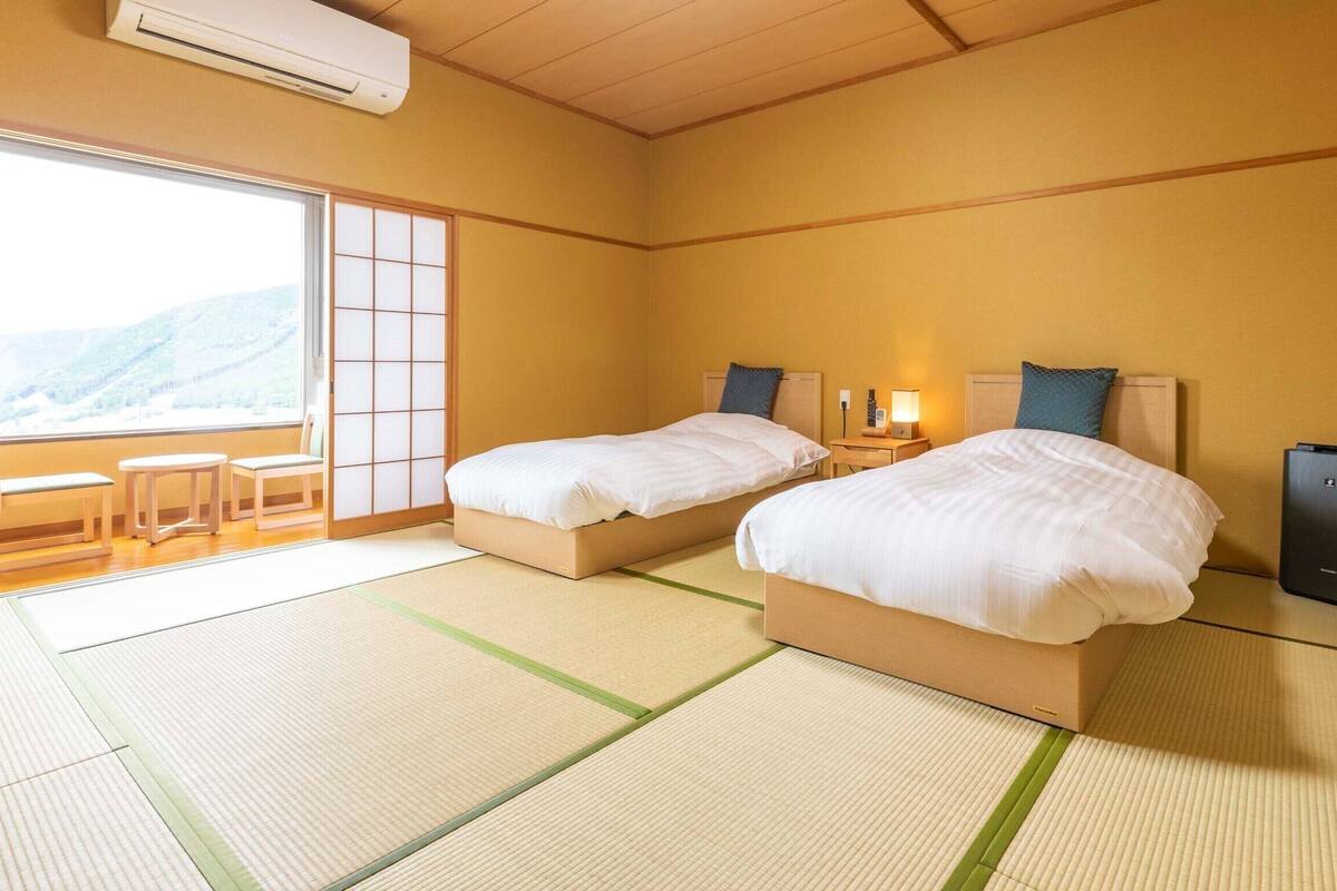Une chambre d’hôtel avec deux lits, une porte coulissante donnant sur un balcon, une table de chevet avec une lampe et un haut-parleur.