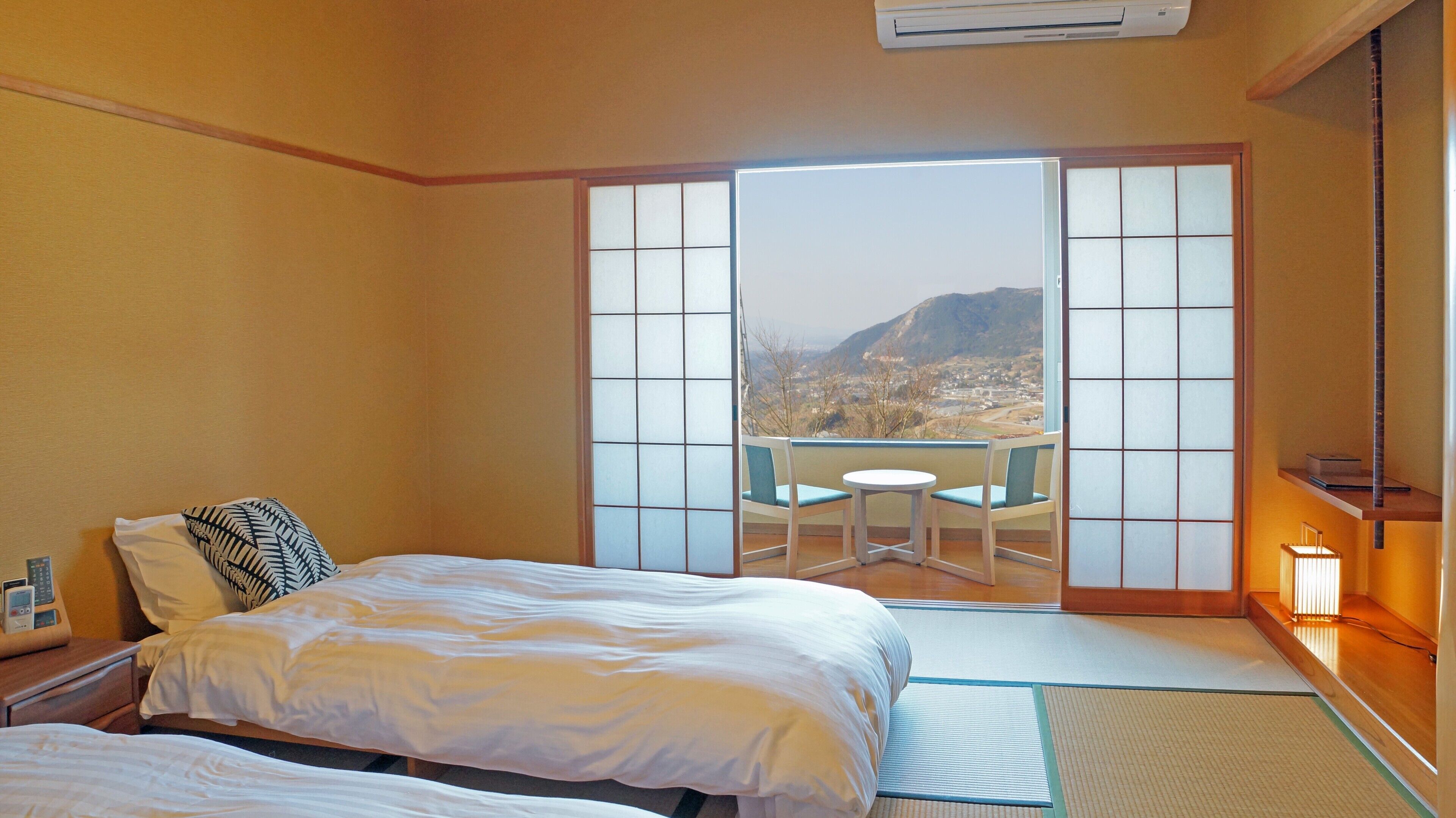 Quarto de hotel com cama, uma pequena mesa com cadeiras, vista para uma paisagem montanhosa através de uma porta deslizante e uma porta deslizante em estilo tradicional japonês.