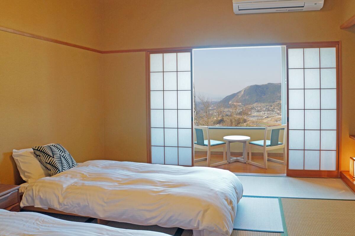 Une chambre d’hôtel avec un lit, une petite table et des chaises, une vue sur un paysage montagneux par une porte coulissante, et une porte coulissante de style japonais traditionnel.