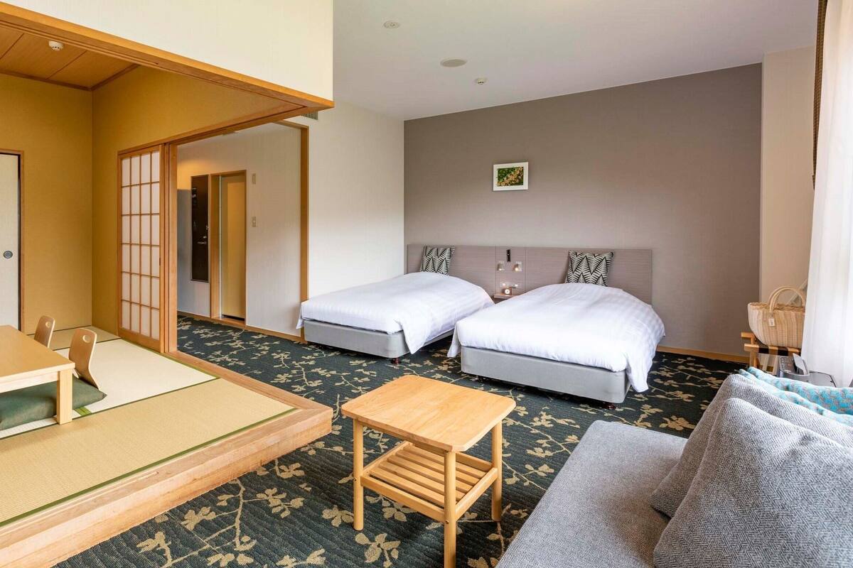 Une chambre d’hôtel avec deux lits, une petite table, un canapé, un sol recouvert de tatamis et un paravent japonais.