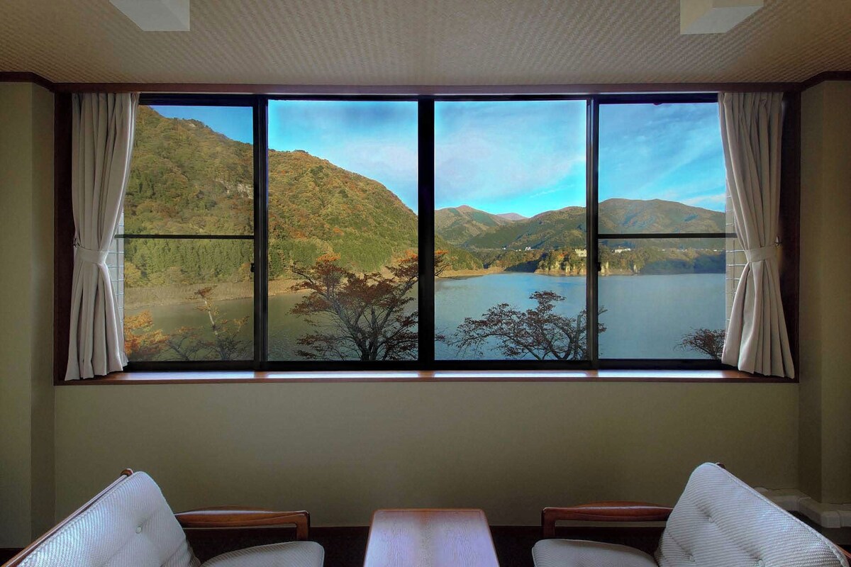 A room with a large window offering a view of a lake and hills. There are two chairs and a small table in the foreground.