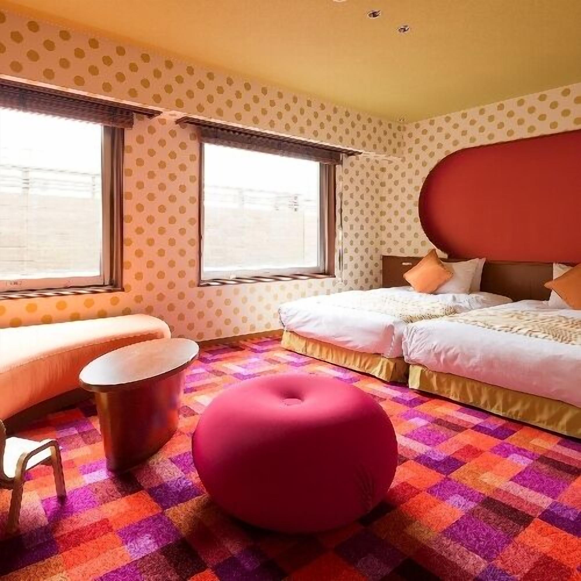 Una camera d'albergo con un letto, un poggiapiedi rosso, un tavolino e una finestra.