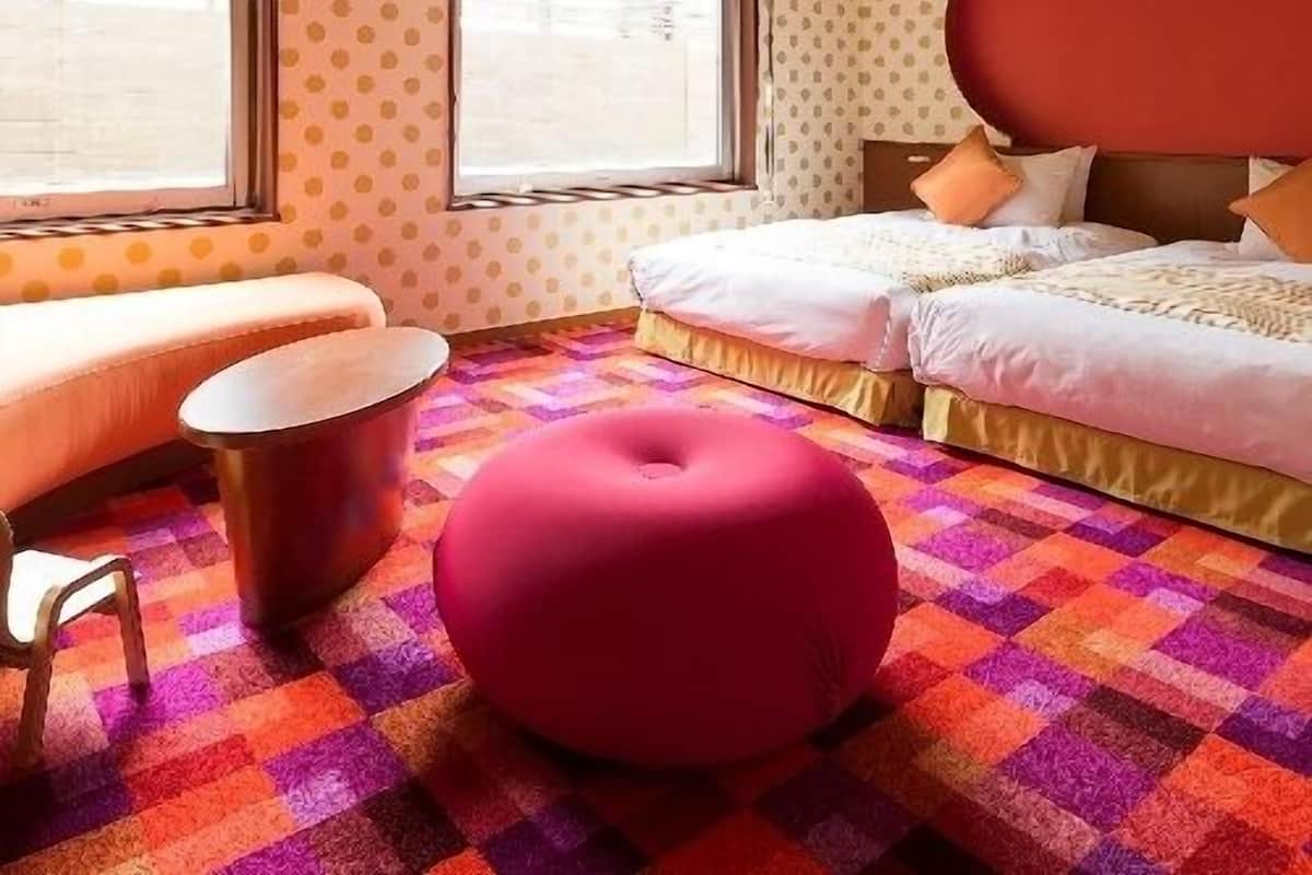 Ein Hotelzimmer mit einem Bett, einem roten Hocker, einem kleinen Tisch und einem Fenster.
