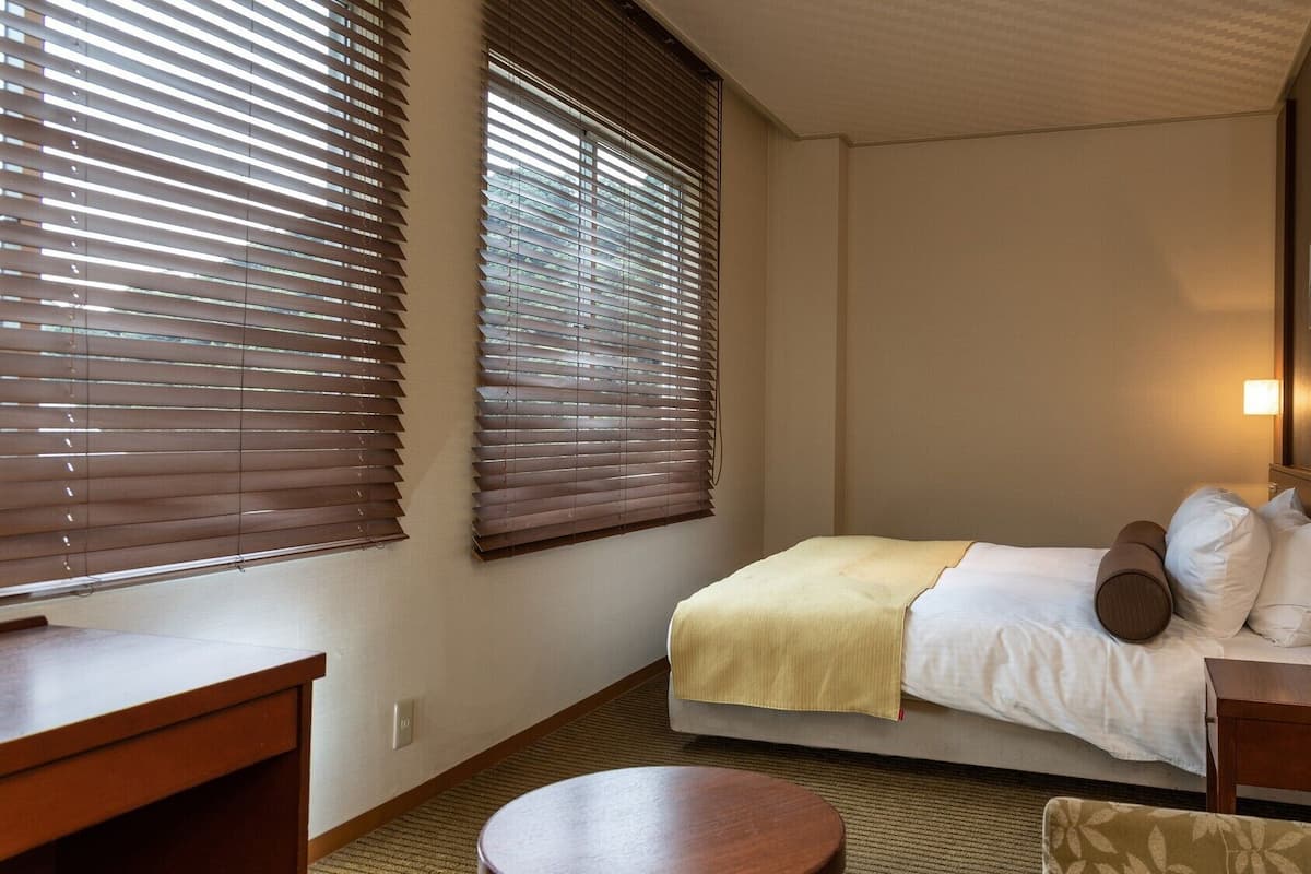 Ein Hotelzimmer mit einem Bett, Nachttischen, einem runden Tisch und zwei Fenstern mit Jalousien.