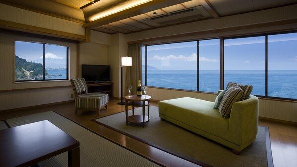 A hotel room with a large window offering a view of the sea and a mountain, a sofa, a chair, a television, and a small table with a bottle and glasses. - Yaizu Grand Hotel (Yaizu)