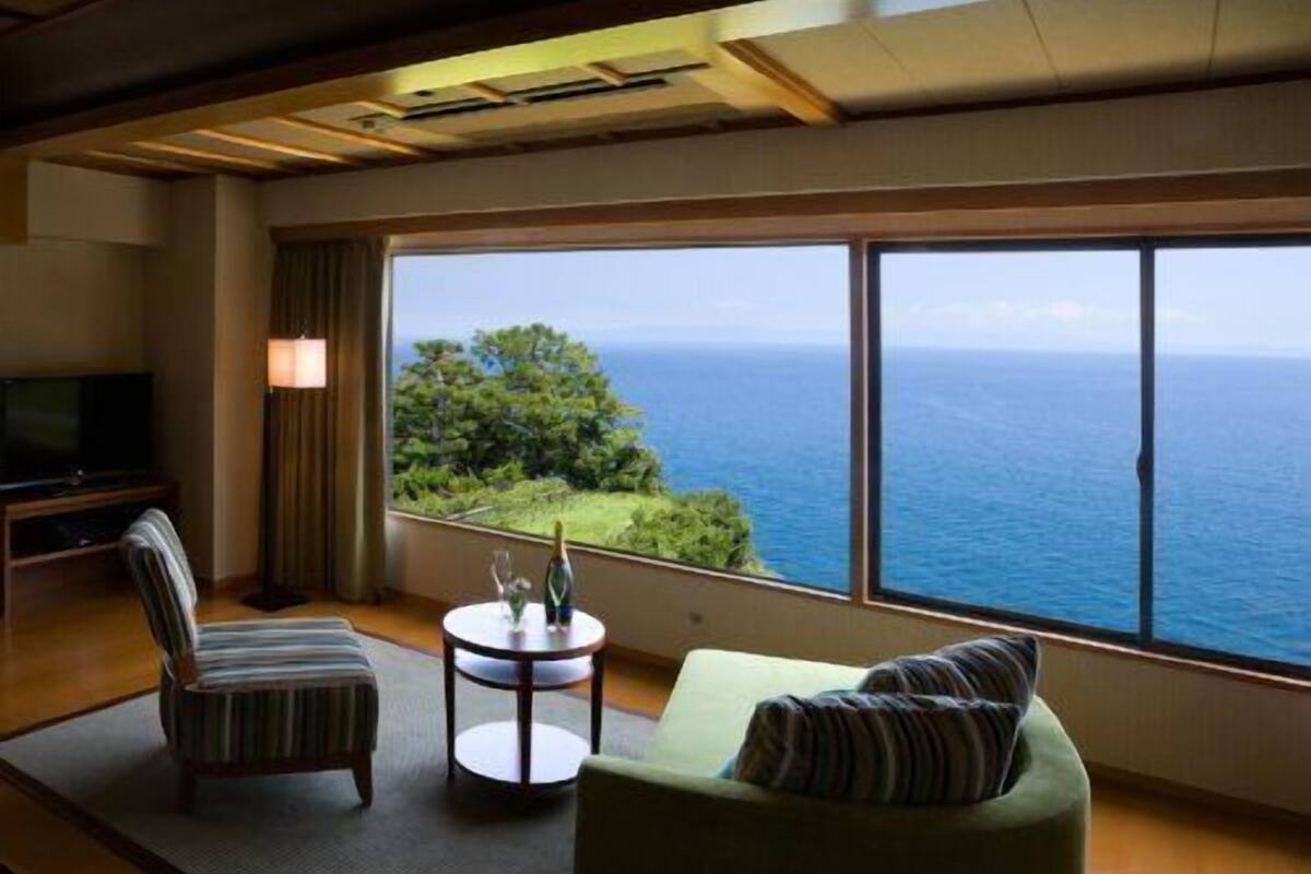 Ein Zimmer mit großem Fenster, einer Couch, einem Sessel, einem kleinen Tisch und einem Fernseher.