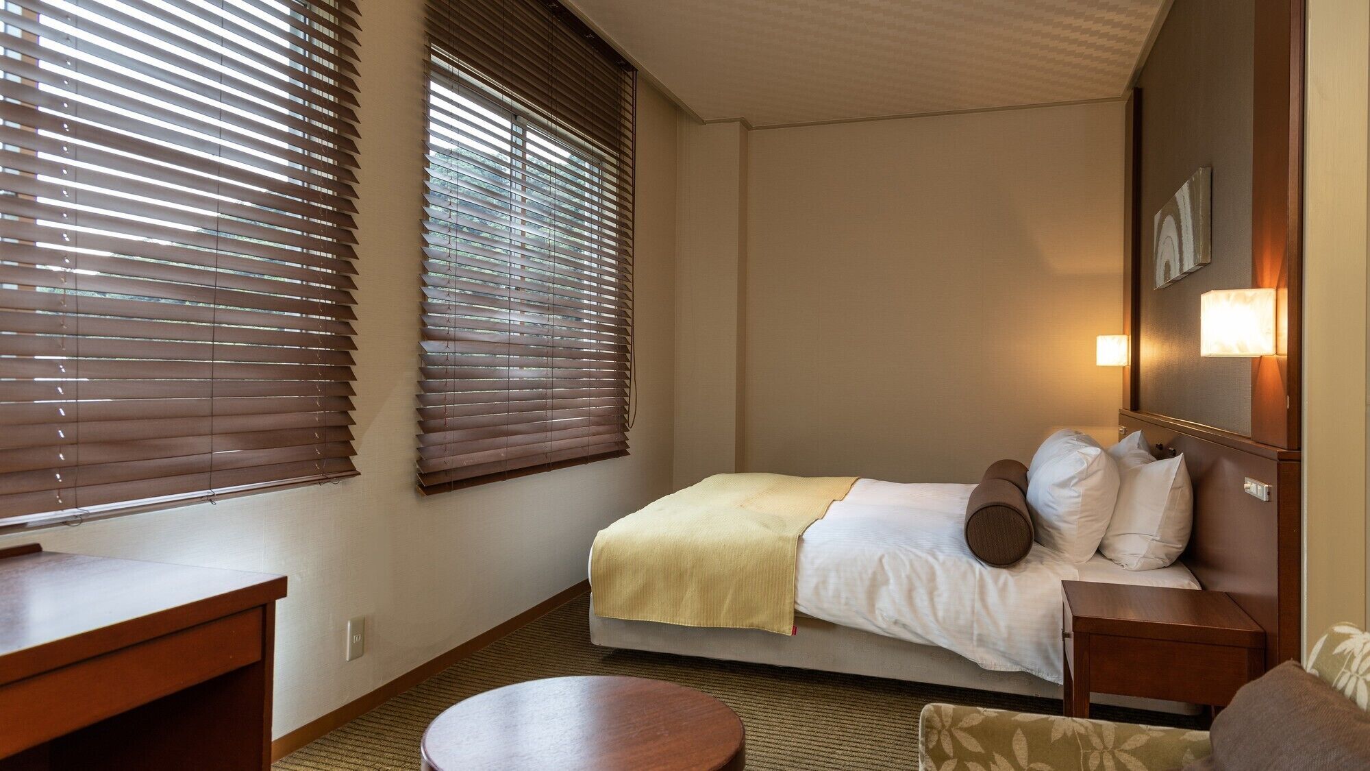 Une chambre d’hôtel comprenant un lit, des tables de chevet, une table ronde et deux fenêtres équipées de stores.
