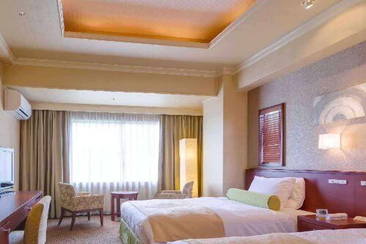 Ein Hotelzimmer mit zwei Betten, einem Schreibtisch, einem Stuhl, einer Lampe und einem Fenster mit Vorhängen.