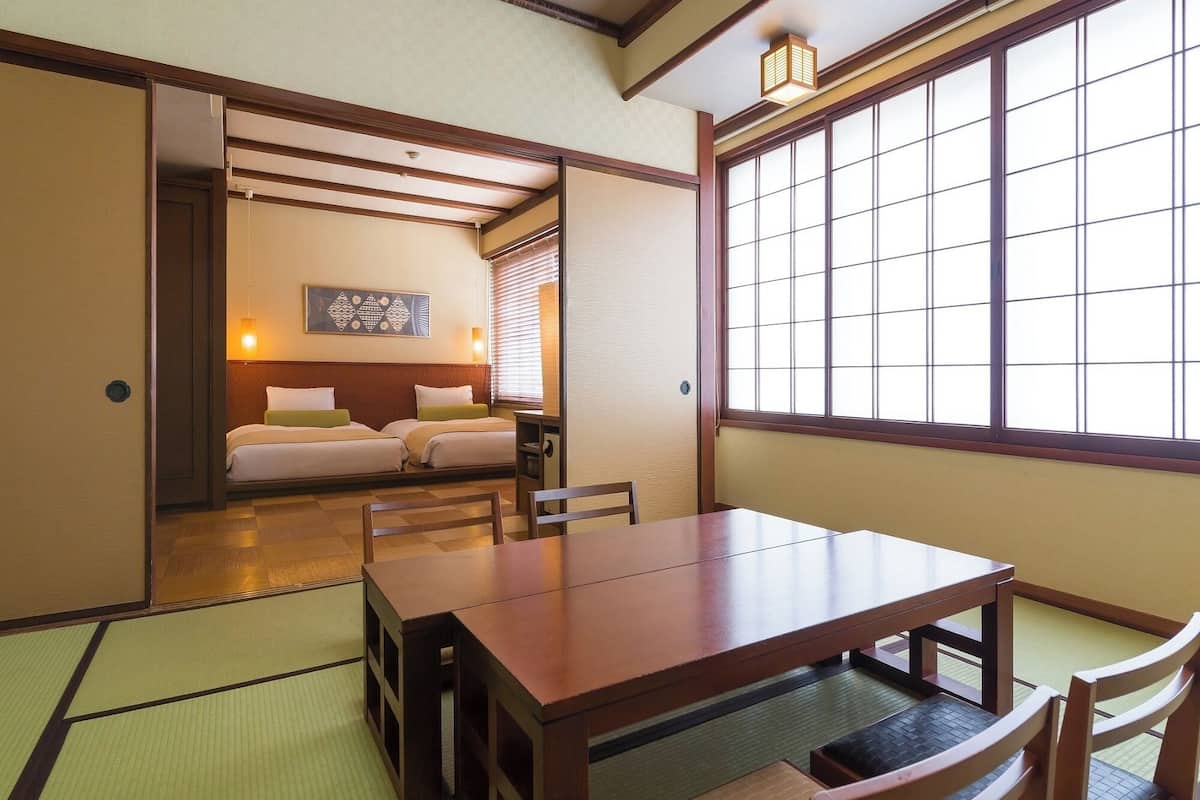 Ein traditioneller japanischer Raum mit Tatami-Böden, einem niedrigen Holztisch und einer verschiebbaren Tür zu einem Schlafzimmer mit zwei Betten.