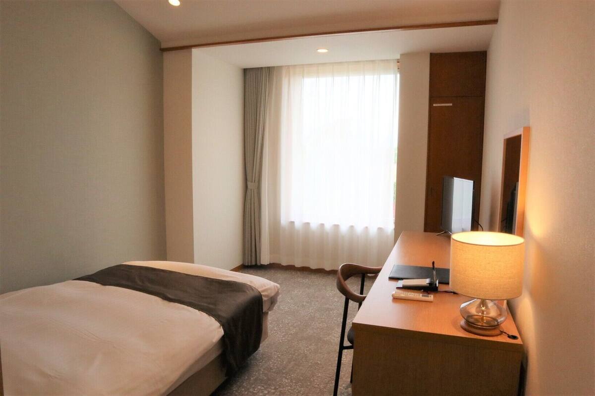 Une chambre d’hôtel avec deux lits, un bureau avec une lampe, une chaise et une grande fenêtre avec des rideaux.