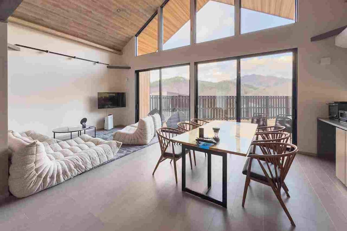 A modern living space with a large dining table, comfortable seating, a flat-screen TV, and a view of the outdoors.