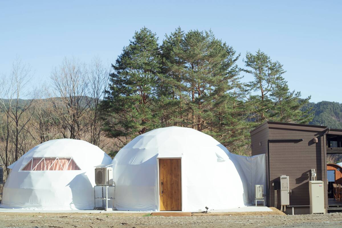 Deux structures en forme de dôme, dotées d’une porte en bois et d’un appareil de climatisation, sont intégrées à un paysage naturel, avec des arbres et un bâtiment en arrière-plan.