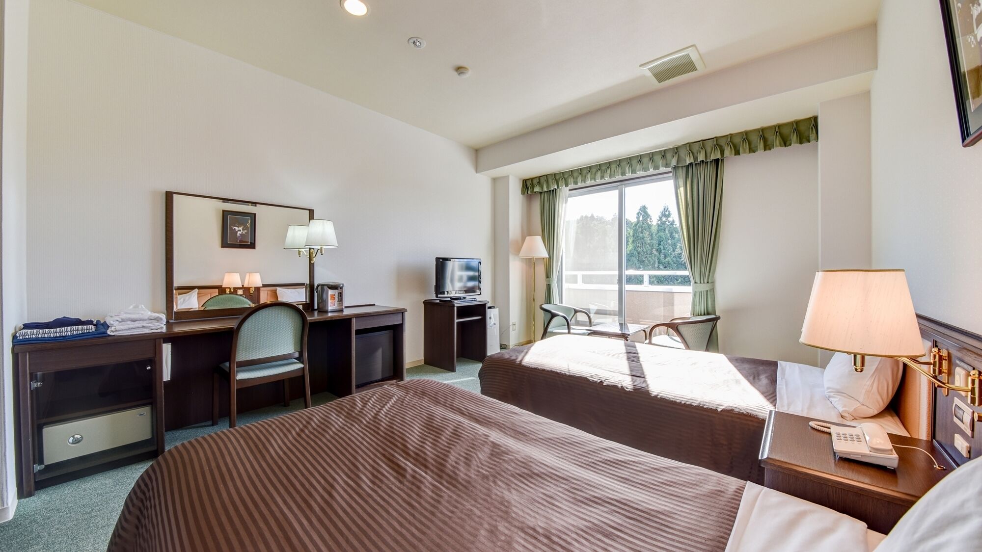 Ein Hotelzimmer mit zwei Betten, einem Schreibtisch mit Stuhl, einem Fernseher, einer Schreibtischlampe und einem Fenster mit Vorhängen.