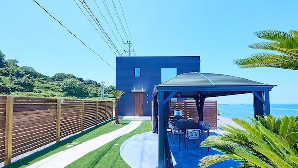 A modern house with a blue exterior, a wooden fence, a patio with a table and chairs, and a view of the ocean.
