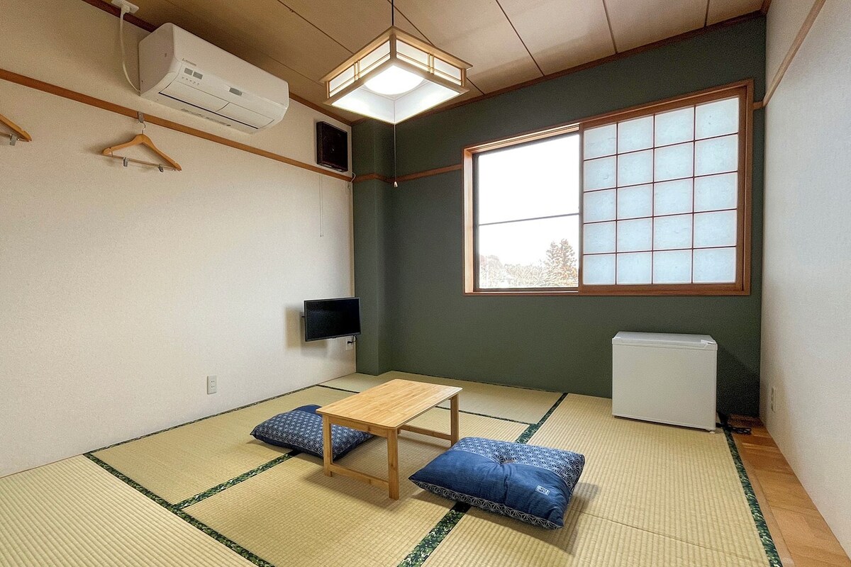 Quarto com piso de tatame, uma mesa de madeira baixa, dois almofados, TV na parede, ar-condicionado e janela com vista.