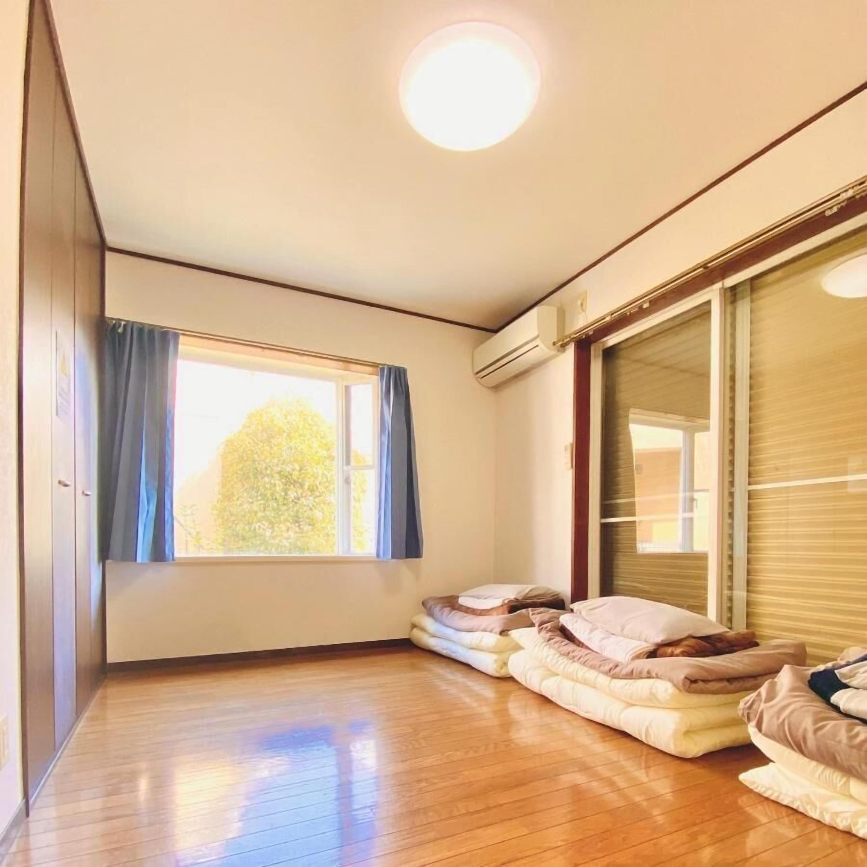 Ein Zimmer mit Holzboden, einem großen Fenster mit Vorhängen, einem Bett mit Kissen und einer Klimaanlage.