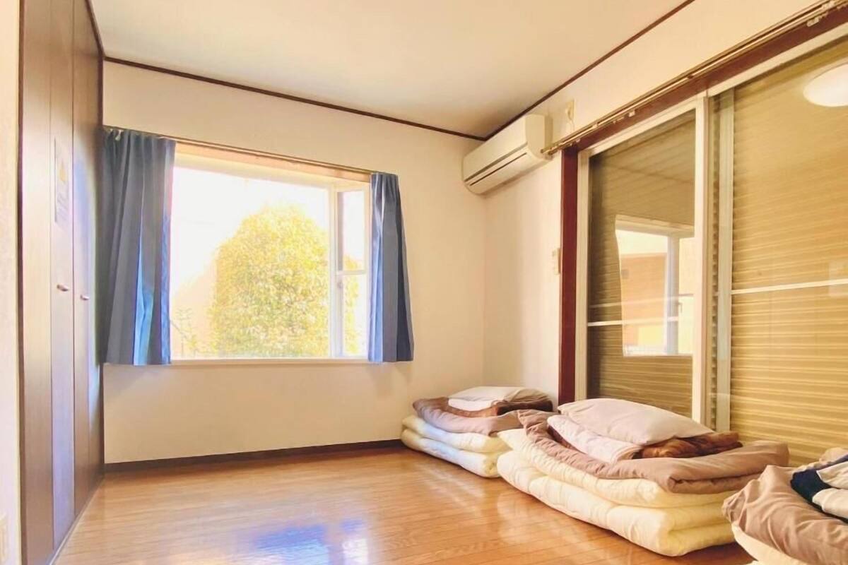 Ein Zimmer mit Holzboden, einem großen Fenster mit Vorhängen, einem Bett mit Kissen und einer Klimaanlage.