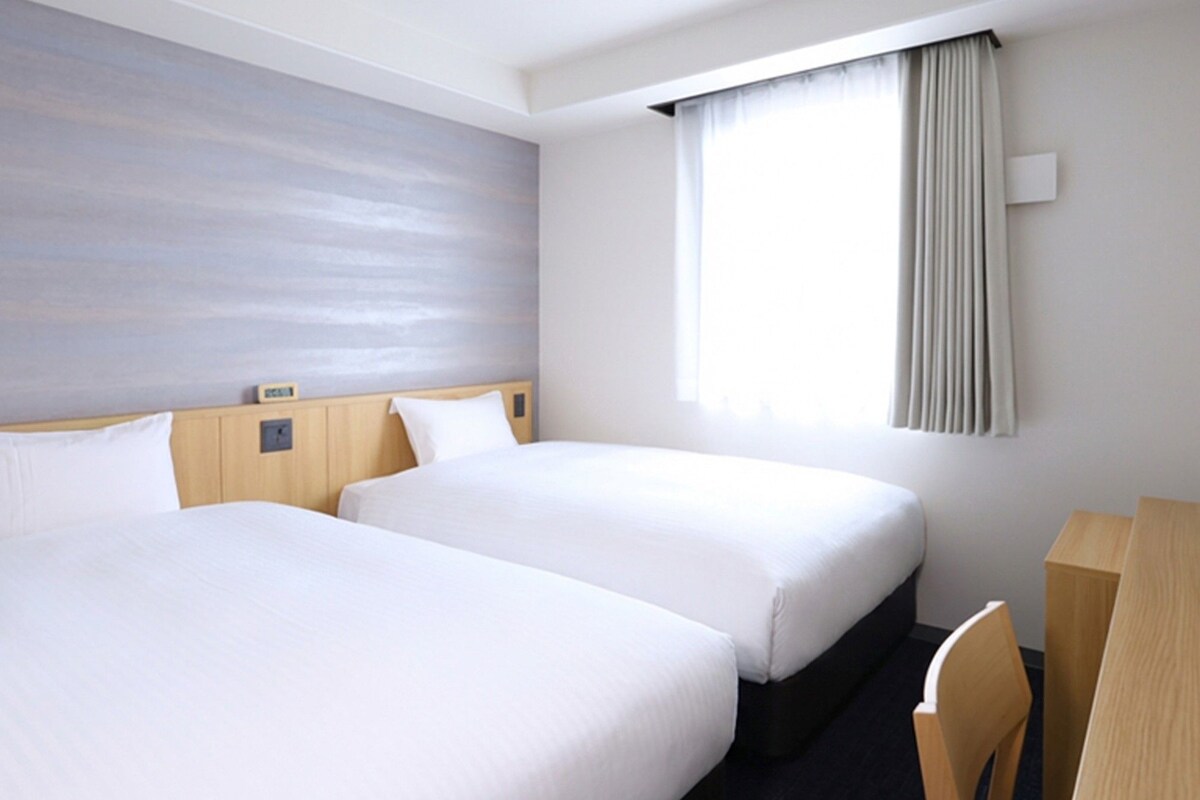 A hotel room with two beds, a desk, a chair, a window with curtains, a wall clock, and a flat-screen TV.