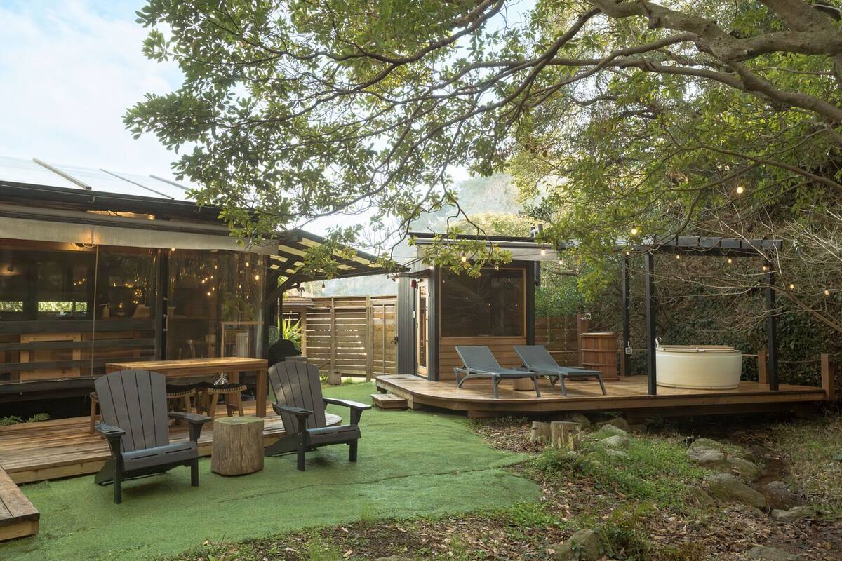 Un patio exterior moderno con terraza de madera, sillones de descanso, bañera de hidromasaje y una estructura con paredes de cristal.