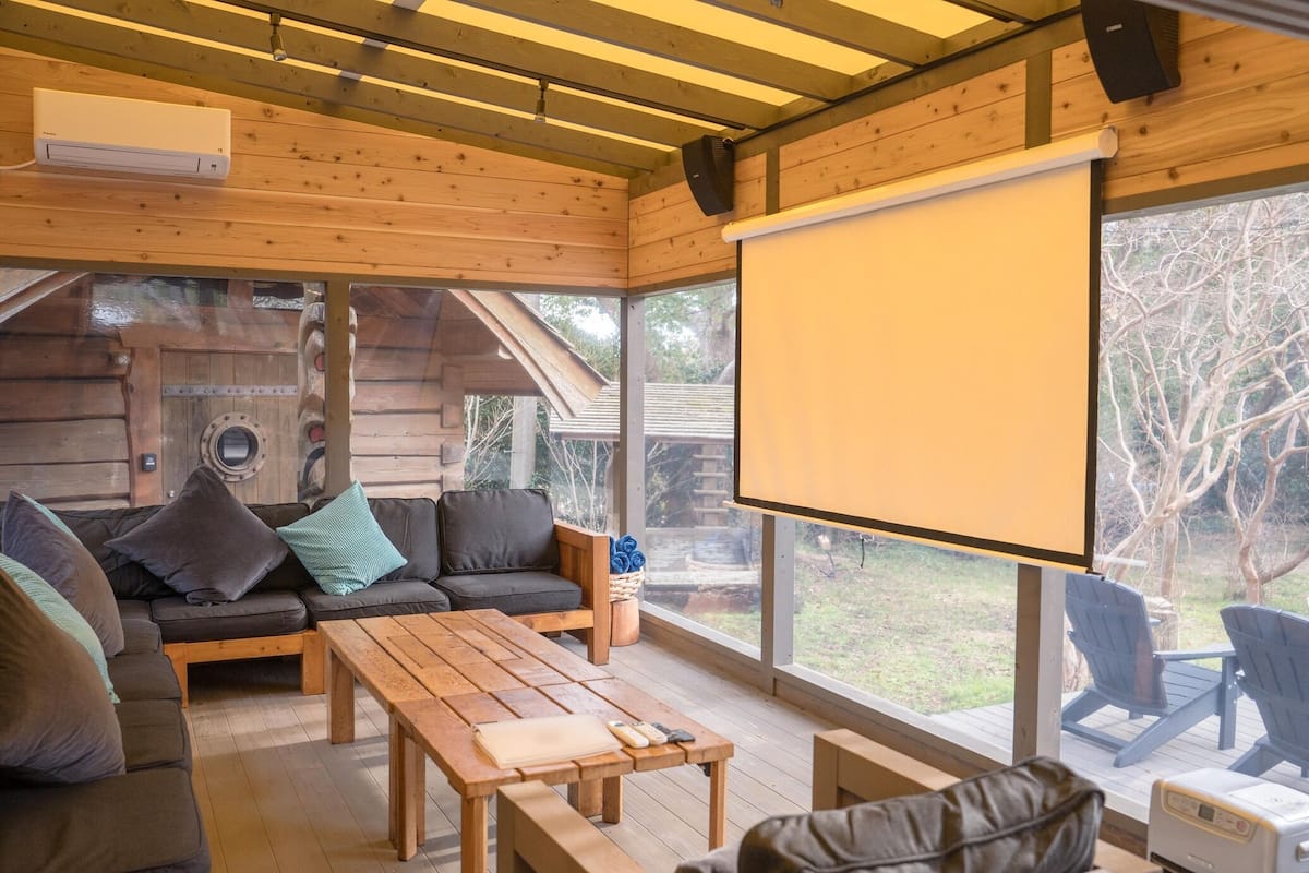 Interior de una cabaña de madera con sofá, mesa, sillas, pantalla de proyector y ventanas amplias.